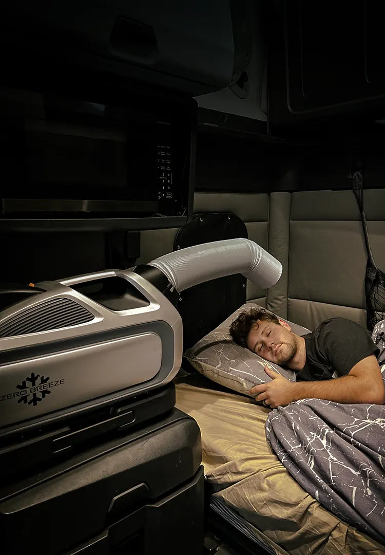 A man sleeps peacefully in a sleeper cab of a truck while the Mark 3 cools the space beside him. The AC unit is positioned on a storage surface with a flexible air duct directed toward the bed.