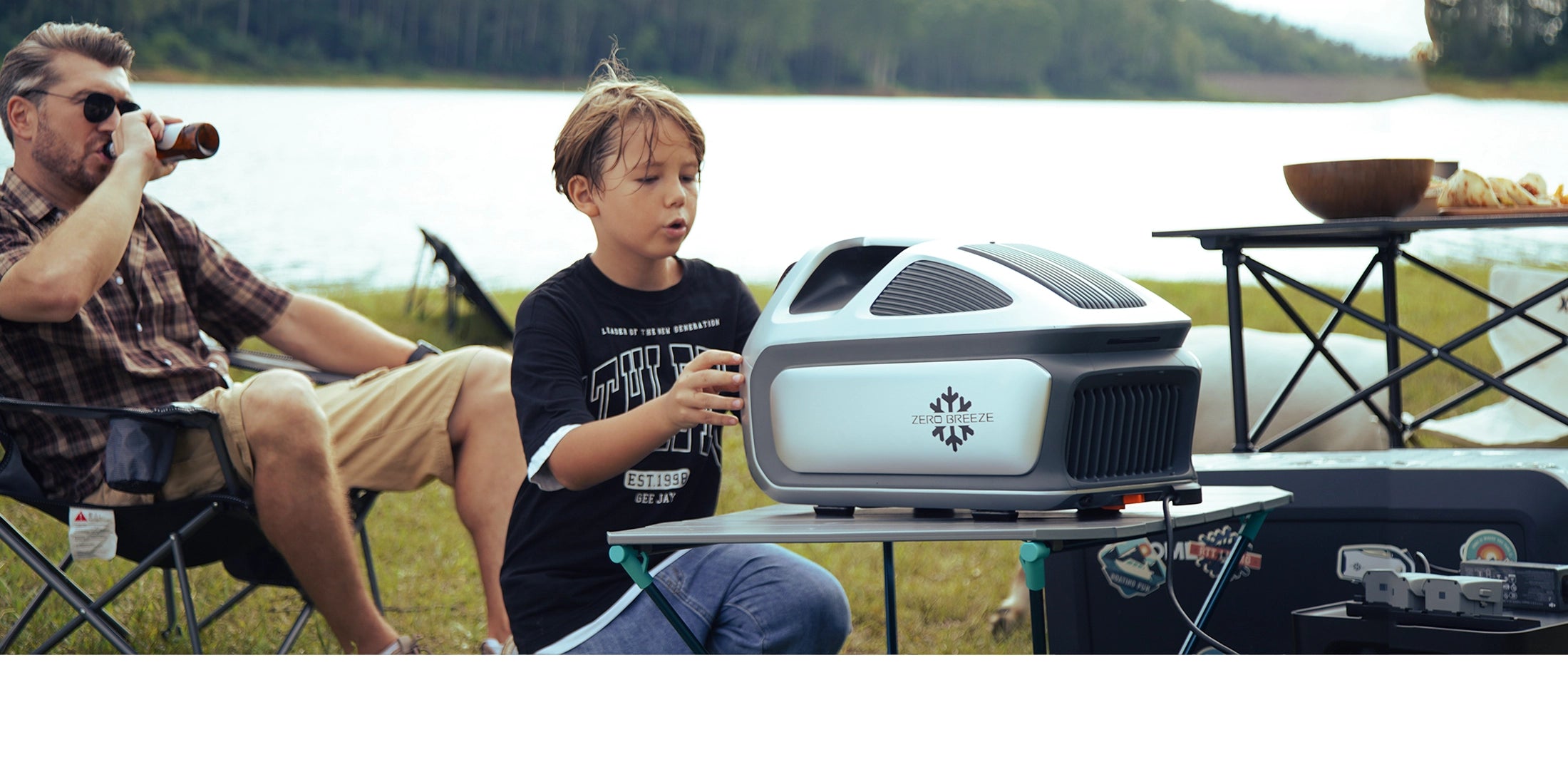 A child interacts with the ZERO BREEZE Mark 3 portable air conditioner at a lakeside camping setup, with adults relaxing nearby and camping gear in the background.