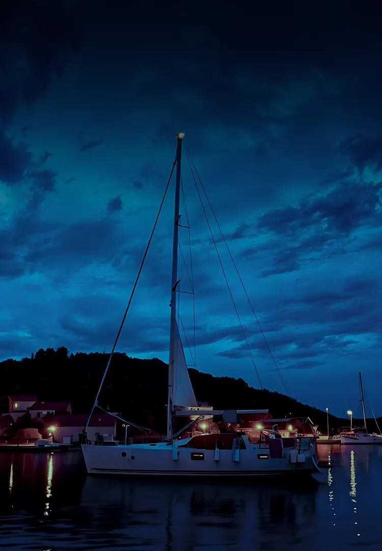 A serene nighttime scene at a marina featuring a white sailboat docked near the shore. The boat is illuminated by warm dock lights reflecting on the calm water, with dark hills and deep blue clouds in the background.