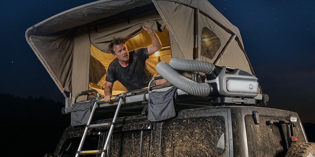 A sweaty man looks out from a rooftop tent at night. A Mark 3 portable AC is mounted on the overland rig's roof, with dual hoses providing cool air to the tent under a starry sky.