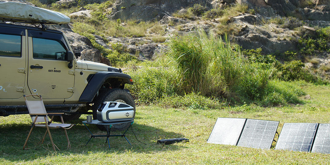 A Mark 3 portable AC sits on a small table next to an off-road jeep. Portable solar panels are laid out on the grass nearby to charge the unit during an outdoor camping trip.