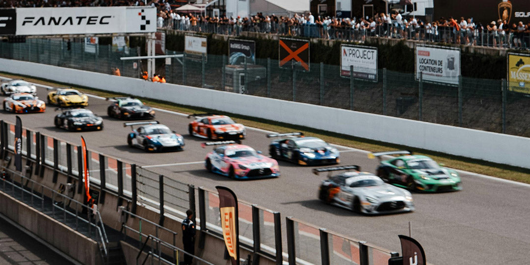 A dynamic scene from a motorsport event shows multiple race cars are speeding down the track, with fans in the background cheering as the cars race past the pit area.