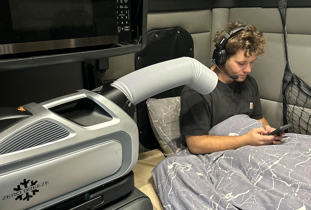 ZERO BREEZE Mark 3 portable air conditioner is in use inside a truck sleeper cabin, cooling a resting man with a front air outlet duct while he uses his phone.