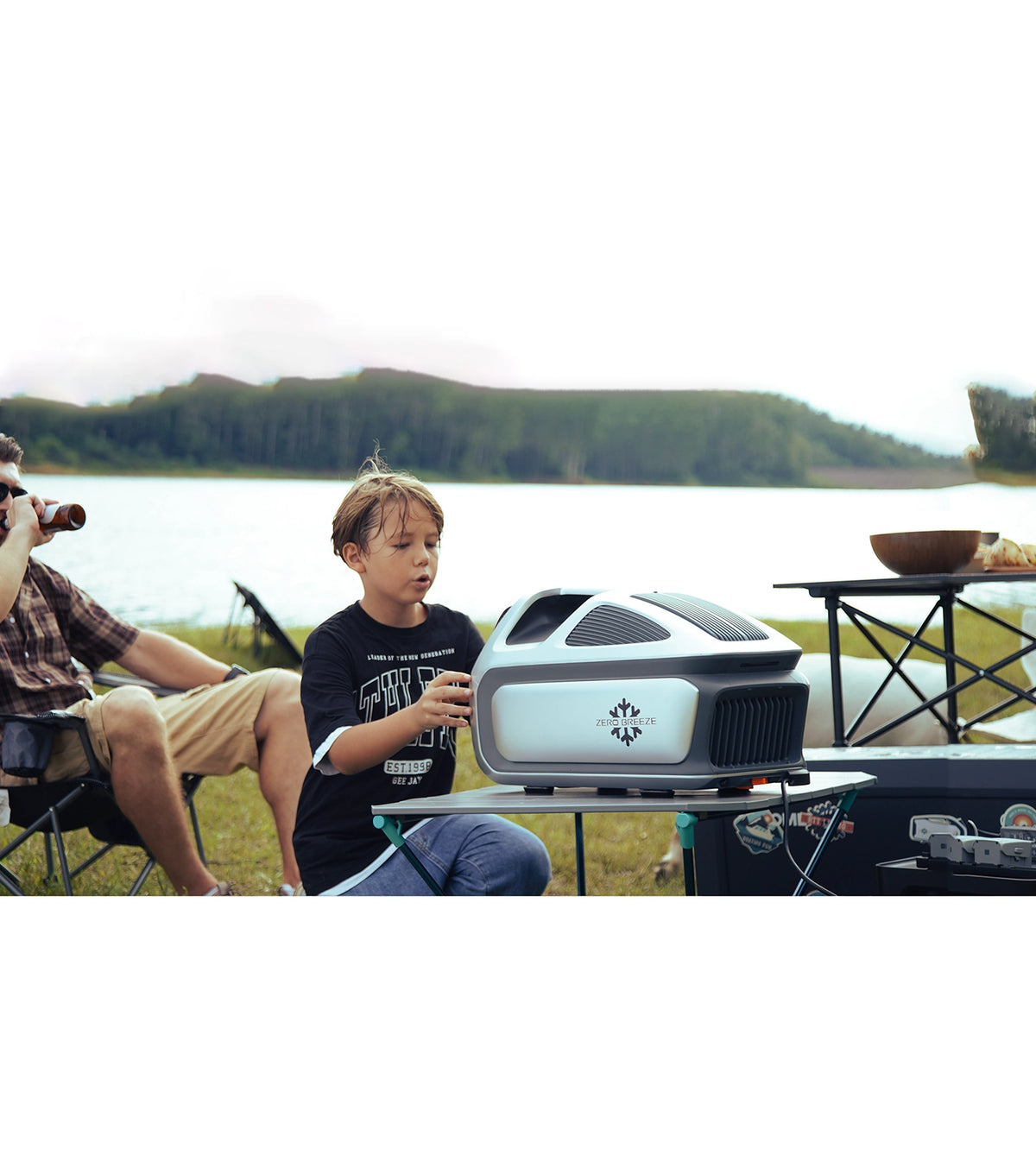 A child interacts with the ZERO BREEZE Mark 3 portable air conditioner at a lakeside camping setup, with adults relaxing nearby and camping gear in the background.