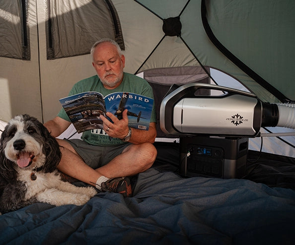 A man reads a magazine inside a tent with his dog beside him and a ZERO BREEZE Mark 3 portable air conditioner running on a power station.