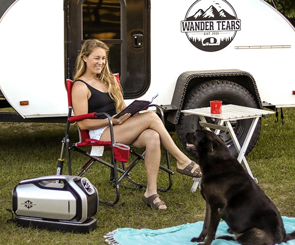 A woman relaxes in a camping chair outside a teardrop trailer with a dog nearby, while the portable air conditioner runs beside her on the grass.