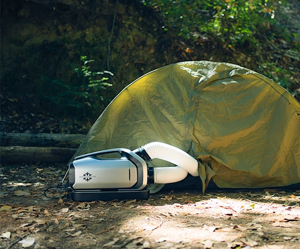 The portable air conditioner is set up outside a small camping tent in the woods, with dual ducts connected for cooling airflow.